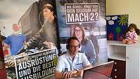 Im Hintergrund Werbeplakate der Bundeswehr. Davor sitzt ein Soldat mit Brille und Headset vor einem Laptop.