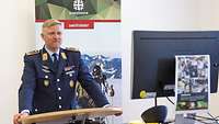 Ein Soldat in dunkelblauer Luftwaffen-Uniform steht an einem Rednerpult. Er spricht in Richtung Monitor.