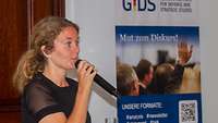 Die Vortragende Dr. Marie-Theres Beumler spricht ins Mikrofon, neben ihr steht ein Plakat vom GIDS