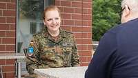 Eine rothaarige Frau in Tarnfleck-Uniform sitzt einem Mann im dunklen Pullover gegenüber. Sie lächelt ihn an.