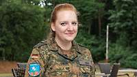 Eine junge rothaarige Frau in Tarnfleck-Uniform. Sie sitzt auf einem Stugl und lächelt in die Kamera.