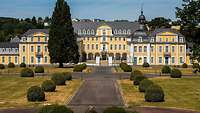 Ein gelbes Schloss mit grauem Dach, davor ein Weg und Büsche.