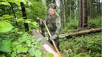 Ein Soldat entfernt mit einem Werkzeug aus Metall die Rinde von einem großen Stamm im Wald.