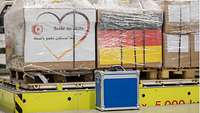 Drei mit Kisten bepackte Transportpaletten, auf der linken ist ein Aufkleber mit einem Herz und dem Schriftzug „Seite an Seite“