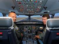 Multinationales Cockpit. Zwei Soldaten sitzen in einem Flugsimulator und schauen über ihre Schultern.