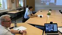 Drei Personen schauen auf die große Leinwand, auf der die Teilnehmer der Videokonferenz zu sehen sind.