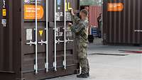 Ein Soldat steht an einem braunen Container und markiert ihn mit gelben Klebestreifen.