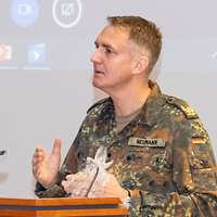 Brigadegeneral Neumann steht am Rednerpult. Im Hintergrund läuft eine Videokonferenz