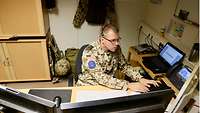 Ein Soldat sitzt am Schreibtisch mit mehreren Monitoren und tippt auf seiner Tastatur, auf dem Kopf trägt er ein Headset