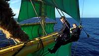 Ein Soldat und eine Soldatin in blauem Overall stehen auf einer Rah eines Masts eines Segelschiffs; im Hintergrund Meer.