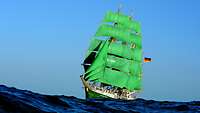 Ein Segelschiff mit grünem Rumpf und grünen Rahsegeln auf dem Meer vor hellblauem Himmel.