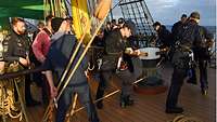 Soldaten in blauer Arbeitsuniform lehnen sich in Holzstangen an einem zylinderförmigen Objekt auf dem Deck eines Segelschiffs.