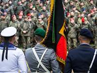 Drei Soldaten unterschiedlicher Teilstreitkräfte stehen in Rückenansicht mit der Truppenfahne vor angetretenen Soldaten.