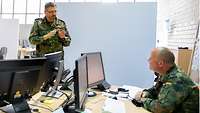 Generalarzt Dr. Backus steht vor einem Schreibtisch und unterhält sich mit einem Soldaten, der zwei Monitore vor sich hat.