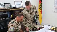 Ein sitzender und ein stehender Soldat blicken in eine Mappe auf einem Schreibtisch und besprechen deren Inhalte