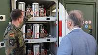 Ein Soldat erklärt dem Ministerpräsidenten die Sauerstofferzeugungsanlage. Beide stehen davor.