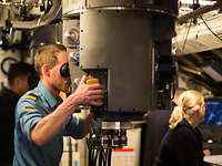 U-Boot WO-Schülerfahrt Periskop Ein Marinesoldat in blauer Arbeitsuniform steht vor einem grauen zylinderförmigen Objekt, das aus der Decke kommt.