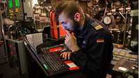 Ein Marinesoldat in blauer Arbeitsuniform sitzt in einem Raum mit Anzeigen und Rohrleitungen, ein Laptop auf den Knien.