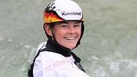 Eine junge Frau mit Helm in Weiß und den Deutschlandfarben sitzt in ihrem Kajak auf dem Wasser und lächelt in die Kamera. 