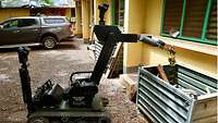 Ein Roboter hält in seinem Greifarm eine selbstgebastelte Spreng- und Brandvorrichtung, im Hintergrund ein Haus und ein Auto 