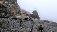 Ein Mann steigt mithilfe von Griff und Trittschlingen einen Felsen im Nebel hinauf.