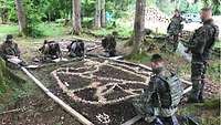 Sieben Soldaten hocken und stehen um einen Geländesandkasten auf dem Waldboden herum.