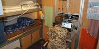 Ein Soldat sitz vor einem Schreibtisch auf seiner Unterkunft an Bord. Links ist ein Doppelstockbett 