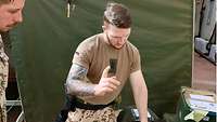 Ein Soldat steht umringt von Gepäck vor einem Zelt und kontrolliert eine Liste