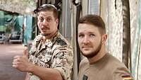 Zwei Soldaten blicken in die Kamera, der Soldat links hält den Daumen hoch, im Hintergrund Häuser