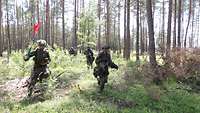 Bewaffnete Soldaten rennen durch den Wald, einer mit roter Fahne, die auf scharfes Feuer hinweist.