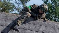 Ein Soldat liegt mit dem Oberkörper auf der Spitze einer hohen Holzwand, mit dem rechten Bein hat er die Wand bereits überwunden