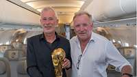 Andreas Brehme und Edgar Ludwigs stehen nebeneinander mit Weltmeisterpokal in der Hand.