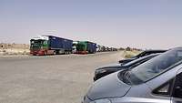 Mehrere mit Containern beladene Sattelschlepper stehen in Doppelreihe entlang einer Straße. Rechts davon befinden sich Pkw