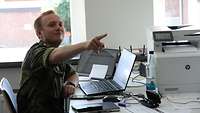 Leutnant Eugen Leikom sitzt am Schreibtisch vor einem PC und zeigt auf etwas mit dem Finger.