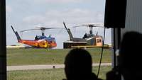 Blick von einer Halle aus: zwei alte Rettungshubschrauber in verschiedenen Farben auf dem Rollfeld.