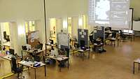 Mehrere Arbeitsplätze sind während Covid-19 in einem großen Saal verteilt. Leinwände stellen ein Lagebild dar.