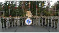 Gruppenbild mit 55 Soldaten und Zivilisten unterschiedlicher Nationen, links ein Wappen, rechts ein Wegweiser