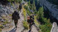 Mehrere Soldaten marschieren mit Ausrüstung einen schmalen Bergpfad hoch. 