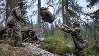 Zwei Soldaten bauen gemeinsam im Wald ihre Zelte auf und werfen einen Rucksack. 