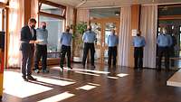 Sieben Männer stehen in einem großen Raum. Ein Mann im Anzug liest sechs Männern in Feuerwehr-Uniform vor.