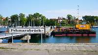 In einem Hafen liegt ein Ponton. Davor das Schild Fehmarnbrücke.