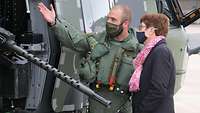 Ein Soldat erklärt einer Frau in Zivilkleidung die lafettierte, aus der Seitentür ragende Bordwaffe eines Hubschraubers.