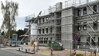 Die Baustelle eines großen Zweckbaus mit Gerüst. Daneben eine hohe Birke.