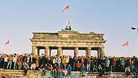 Viele Menschen, die auf und vor der Berliner Mauer stehen. Im Hintergrund steht das Brandenburger Tor.