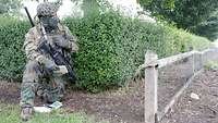 Ein Soldat kniet, das Funkgerät auf dem Rücken, sein Gewehr vor der Brust, vor einer Hecke und spricht in das Funkgerät.