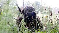 Im hohen Gras liegt ein Soldat. Auf dem Rücken ein Funkgerät, dessen Antennen über ihm zu sehen sind.