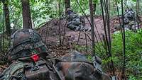 Zwei Soldaten liegen im Wald auf einer Anhöhe. Ein weiterer Soldat bewegt sich von hinten auf sie zu.