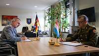 Dr. Dupont und Dr. Baumgärtner sitzen sich mit Abstand an einem Tisch gegenüber und unterschreiben die Dokumente