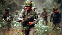 Soldaten in olivgrüner Uniform laufen mit Waffe und Tarnung durch den Wald.