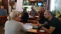 Frauen und Männer sitzen in kleinen Gruppen in einem Café an Tischen und unterhalten sich.